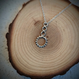 Puede incluir: Un collar con colgante de plata con una cadena y un colgante. El colgante tiene tres pequeños círculos abiertos conectados a un círculo más grande y texturizado. El collar se muestra sobre una superficie de madera.