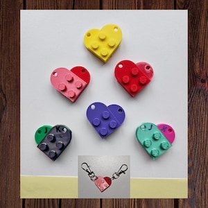May include: Six heart-shaped keychains made of plastic building blocks. Each keychain is a different color: yellow, pink, red, purple, green, and blue. The keychains are arranged in a circle on a white background. A close-up of a pink heart-shaped keychain with a silver chain is shown in the bottom right corner.