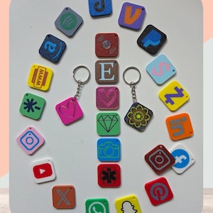 Puede incluir: Una colección de llaveros y colgantes cuadrados de colores, cada uno con un icono o letra diferente. Los llaveros incluyen logotipos de redes sociales, una nota musical y un corazón. La letra "E" también está presente. Los llaveros están dispuestos sobre una superficie blanca.