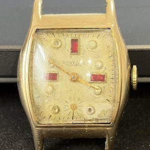 May include: A gold-tone Sanford 17 jewels wristwatch case with a textured off-white dial. The face features rectangular red markers at the twelve, three, and nine o'clock positions, circular markers for other hours, and a small sub-dial at the bottom.