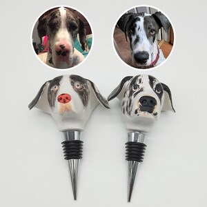 Puede incluir: Dos tapones de botella de vino de cer&aacute;mica con forma de cabeza de perro Gran Dan&eacute;s. Los tapones son blancos con marcas negras y marrones. Los tapones tienen una base de metal plateado y un tap&oacute;n de goma negro.