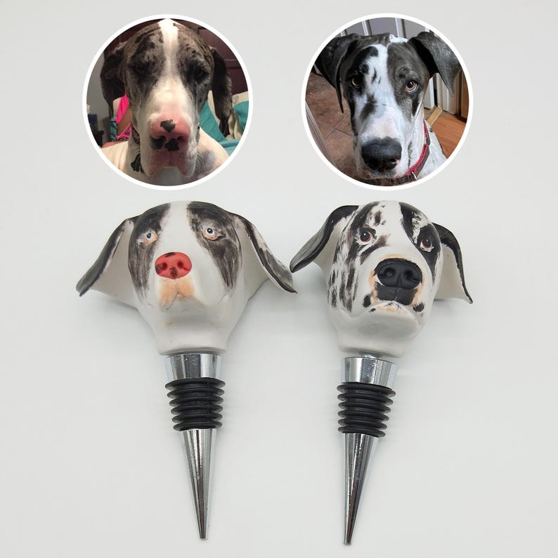 Puede incluir: Dos tapones de botella de vino de cer&aacute;mica con forma de cabeza de perro Gran Dan&eacute;s. Los tapones son blancos con marcas negras y marrones. Los tapones tienen una base de metal plateado y un tap&oacute;n de goma negro.