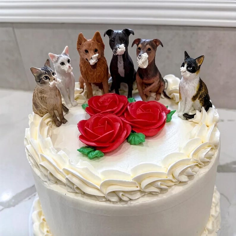 May include: A white frosted cake decorated with red roses and figurines of cats and dogs. The cake has a ruffled edge and is topped with several animal figurines, including a brown tabby cat, a white cat, and several dogs.
