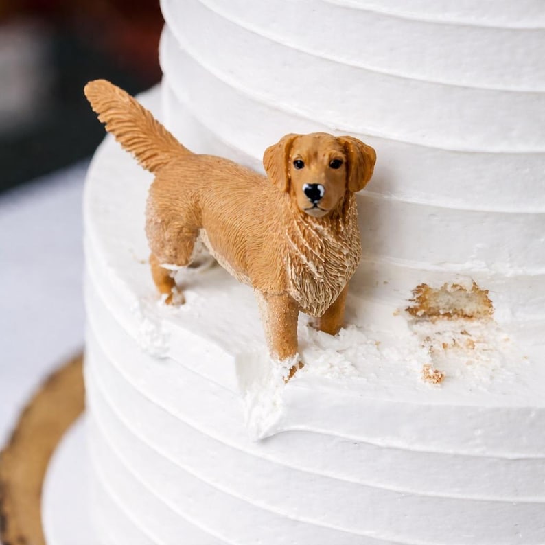 May include: A golden retriever cake topper figurine, standing on a white frosted cake. The dog is a light brown color with detailed fur and features. The cake has a textured, layered design.