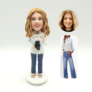 May include: A custom bobblehead figurine of a woman with blonde curly hair, wearing a white blouse, blue jeans, and holding a black camera. The figurine is on a white base with text. Additional images of the woman are included.