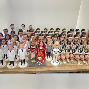 May include: A collection of custom bobblehead figures displayed on a wooden surface. The group includes various individuals in professional attire, sports uniforms, and formal wedding clothing, alongside several detached bobblehead pieces.