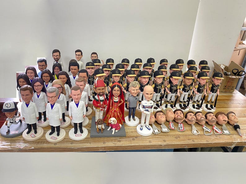 May include: A collection of custom bobblehead figures displayed on a wooden surface. The group includes various individuals in professional attire, sports uniforms, and formal wedding clothing, alongside several detached bobblehead pieces.