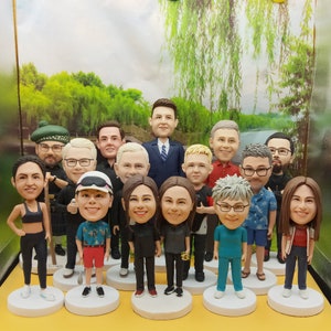 May include: A collection of bobblehead figurines, each depicting a person with unique features and attire. The figurines stand on white circular bases, showcasing a variety of outfits and expressions. The background features a scenic landscape.