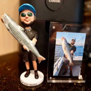 May include: A custom bobblehead figure of a person wearing a blue cap, sunglasses, black t-shirt, and shorts, holding a large silver fish. The circular white base features the text "SO FIN-TASTIC". Next to it is a framed photograph of the same person.