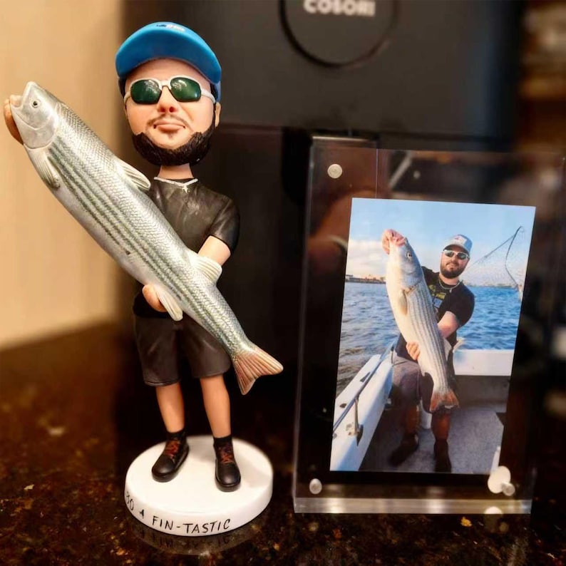 May include: A custom bobblehead figure of a person wearing a blue cap, sunglasses, black t-shirt, and shorts, holding a large silver fish. The circular white base features the text "SO FIN-TASTIC". Next to it is a framed photograph of the same person.