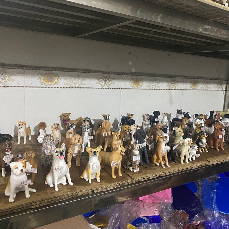 May include: A collection of dog figurines in various breeds and poses, displayed on a shelf. The figurines are painted in realistic colors and have price tags attached. The shelf is made of metal and is in a warehouse setting.