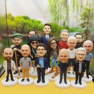 Könnte beinhalten: Eine Gruppe von 14 maßgefertigten Figuren, jede mit einzigartigen Gesichtszügen und Kleidung. Die Figuren stehen auf einem Tisch mit einer gelben Oberfläche. Die Figuren sind alle unterschiedlich groß und in einem Halbkreis angeordnet.