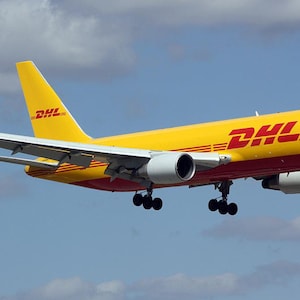 May include: A yellow DHL cargo airplane in flight against a blue sky with clouds. The plane has the DHL logo in red and yellow on the tail and fuselage. The plane's engines and landing gear are visible.