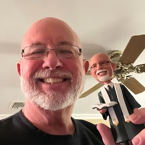 May include: A bobblehead figurine depicting a bald individual with a white beard and glasses, wearing a black robe with a white stole featuring small gold cross emblems, holding an open book in their hands, matching the appearance of the person.