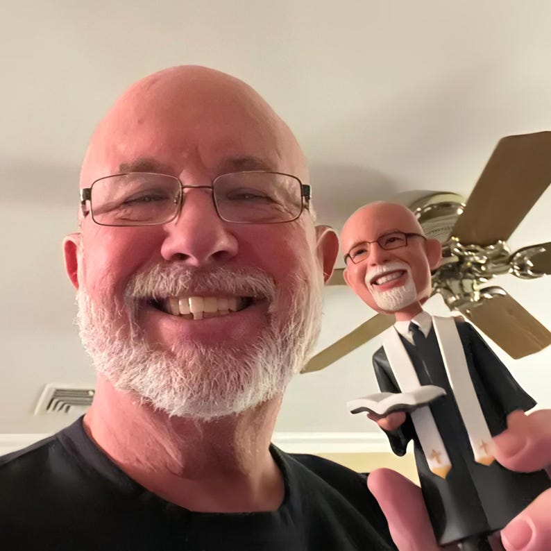 May include: A bobblehead figurine depicting a bald individual with a white beard and glasses, wearing a black robe with a white stole featuring small gold cross emblems, holding an open book in their hands, matching the appearance of the person.