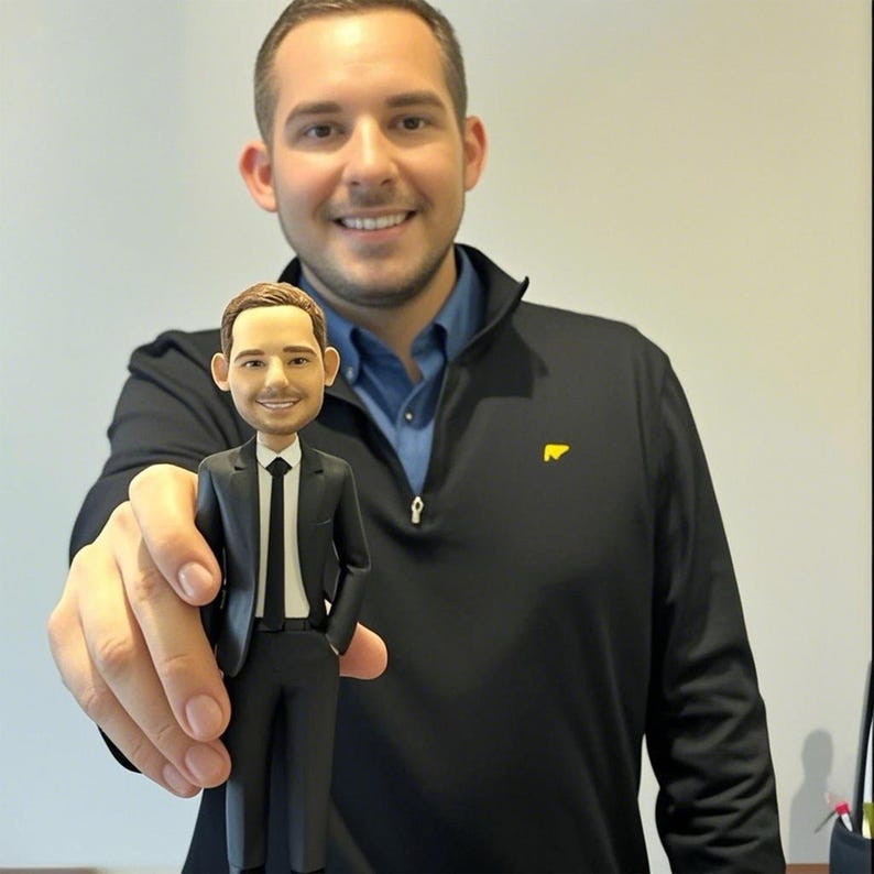 May include: A custom bobblehead figurine depicting a person with short brown hair, wearing a black suit, a white collared shirt, and a black necktie. The figurine features a smiling facial expression and is crafted with a smooth, matte finish.