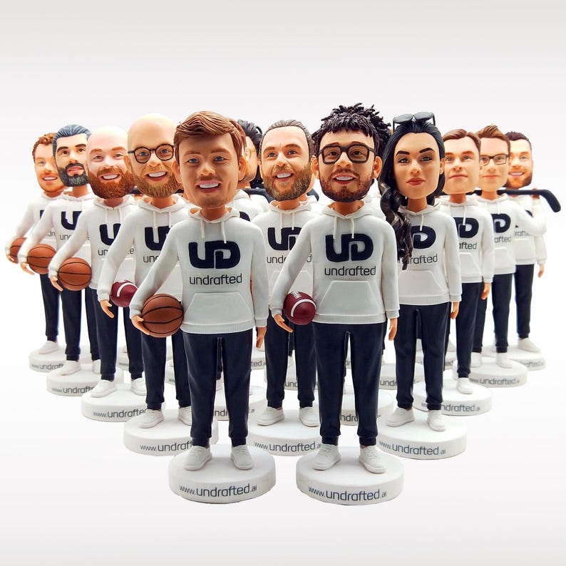 May include: A collection of custom bobblehead figurines, each wearing a white hoodie with the navy blue text "UD undrafted" and dark trousers. Several figures hold miniature brown footballs, while others stand with arms at their sides. Each figure is mounted on a circular white base labelled with the website "www.undrafted.ai".