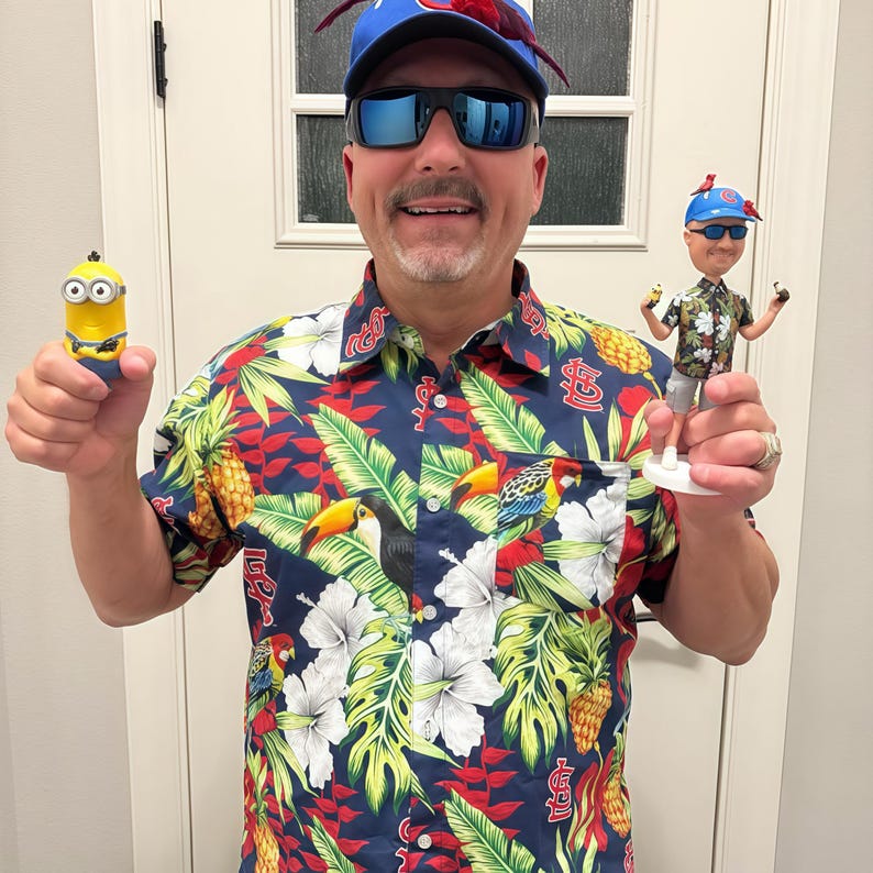 May include: A short-sleeved button-down shirt featuring a navy blue background with a tropical print of pineapples, toucans, parrots, and white hibiscus flowers, accented with red St. Louis Cardinals logos throughout the fabric pattern.