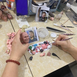 Puede incluir: Una persona est&aacute; esculpiendo un busto de arcilla de la cara de un hombre. El busto est&aacute; sobre una mesa con una variedad de herramientas y materiales, incluyendo una paleta de arcilla de colores, un pincel y un peque&ntilde;o recipiente de pintura blanca.