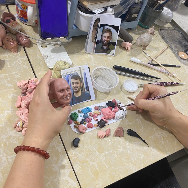 Puede incluir: Una persona est&aacute; esculpiendo un busto de arcilla de la cara de un hombre. El busto est&aacute; sobre una mesa con una variedad de herramientas y materiales, incluyendo una paleta de arcilla de colores, un pincel y un peque&ntilde;o recipiente de pintura blanca.