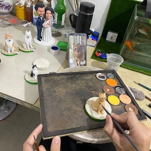 May include: A person is painting a small, tan cat figurine with a paintbrush. Several other figurines, including a wedding cake topper and dog figures, are visible. The figurines are on small, round bases. A smartphone displays a cat image.