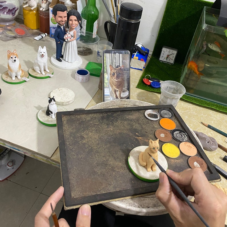 May include: A person is painting a small, tan cat figurine with a paintbrush. Several other figurines, including a wedding cake topper and dog figures, are visible. The figurines are on small, round bases. A smartphone displays a cat image.
