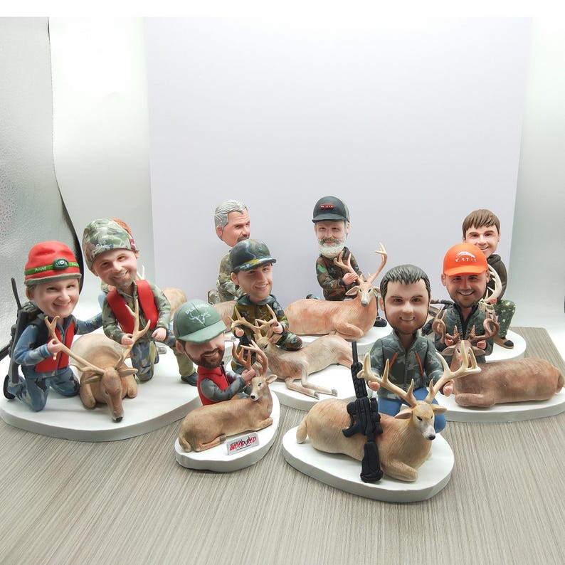 May include: A collection of seven custom bobblehead figurines depicting individuals in hunting gear, each posed with a deer. The figures wear various hats, vests, and camouflage clothing, with some holding rifles, all mounted on individual white oval bases.