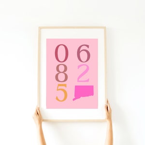 May include: A framed print with a pink background and a white border. The print features a vertical arrangement of numbers in different colours: 06, 82, and 5. The number 5 is orange, the numbers 0 and 6 are pink, and the numbers 8 and 2 are red. The print also includes a pink outline of the state of Connecticut.