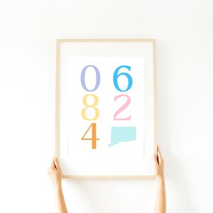 以下が含まれることがあります： 白い背景にカラフルな数字が書かれた額装されたプリント。数字は縦に並べられており、一番上の数字は06、その下に82、そして4と続きます。数字は青、黄色、ピンク、オレンジと異なる色で書かれています。プリントの下部には、コネチカット州の薄い青色の輪郭が描かれています。