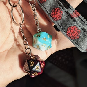 May include: Two dice charms on a silver chain. The dice are both 20-sided and have gold numbers. One die is blue and white, the other is black and red.