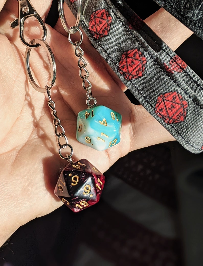 May include: Two dice charms on a silver chain. The dice are both 20-sided and have gold numbers. One die is blue and white, the other is black and red.