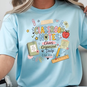 Op de afbeelding: Een lichtblauw t-shirt met een grafische print met de tekst "CLASSROOM NOTES Chaos Organized Daily Est. 2026". Het ontwerp bevat illustraties van een spiraalblok, een geel potlood, een liniaal, een appel en diverse paperclips.