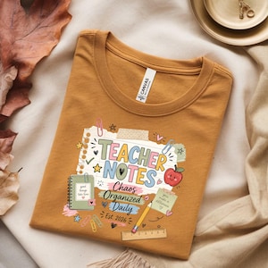 May include: A mustard yellow t-shirt featuring a graphic print of a notebook page with the text Teacher Notes, Chaos, Organized, Daily, and Est. 2026. The design includes illustrations of a spiral notebook, a red apple, a pencil, and a ruler.