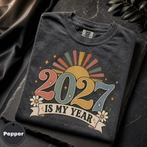 Op de afbeelding: Een antracietgrijs t-shirt met een grafische opdruk met de tekst 2027 IS MY YEAR en GOOD THINGS AHEAD. Het ontwerp bevat een gestileerde zon, kleurrijke cijfers en witte madeliefjes met groene bladeren op de voorzijde van het kledingstuk.