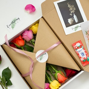 May include: A white box containing a brown paper envelope. The envelope is tied with a pink ribbon and has a round sticker with the text "Earthling Floral" on it. The box contains a bouquet of colourful roses, a card, and a small packet of flower food.
