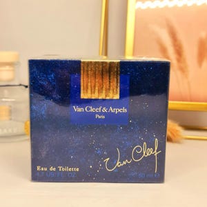 May include: A square perfume box in deep blue with a starry night pattern. It features a gold foil seal at the top center, the text Van Cleef & Arpels Paris, and the signature Van Cleef in gold script. The label reads Eau de Toilette 1.7 US FL OZ.