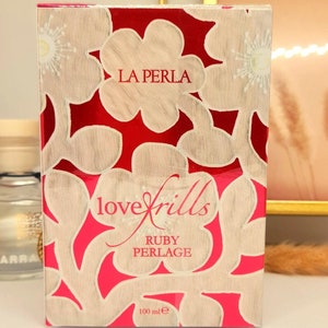 May include: A red box with white floral designs and the text "LA PERLA lovefrills RUBY PERLAGE 100 ml e" on it.