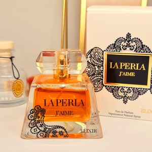 May include: A clear glass bottle of perfume with a gold cap and a black and white lace design. The bottle is labeled "La Perla J'aime Elixir".