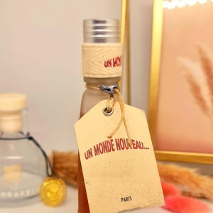 Pu&ograve; includere: Una piccola bottiglia di vetro con tappo argentato e una fascia beige intrecciata attorno al collo. Un'etichetta beige con il testo "UN MONDE NOUVEAU... PARIS" &egrave; attaccata. La bottiglia contiene un liquido marrone chiaro.