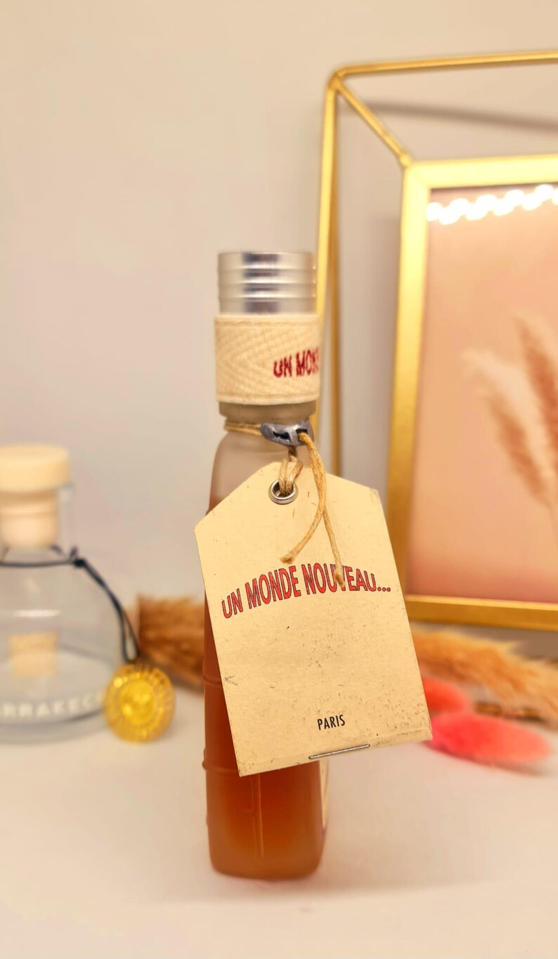 Pu&ograve; includere: Una piccola bottiglia di vetro con tappo argentato e una fascia beige intrecciata attorno al collo. Un'etichetta beige con il testo "UN MONDE NOUVEAU... PARIS" &egrave; attaccata. La bottiglia contiene un liquido marrone chiaro.