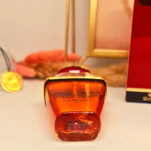 Może przedstawiać: Pomarańczowy flakon perfum ze złotymi akcentami. Butelka ma zaokrąglony kształt i złotą nakrętkę. Etykieta głosi "GUERLAIN PARIS" i "50ml 1.7 FL.OZ". Tło obejmuje czerwone pudełko i złotą ramę.