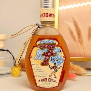 Pu&ograve; includere: Un flacone di profumo con tappo argentato ed etichetta con testo "7 Ciel Heaven" e "Un Monde Nouveau". Il flacone contiene un liquido marrone chiaro e un'etichetta beige con design decorativo.