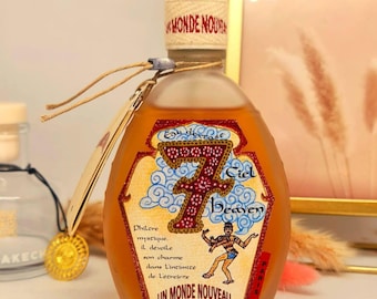 May include: A perfume bottle with a silver cap and a label featuring the text "7 Ciel Heaven" and "Un Monde Nouveau". The bottle has a light brown liquid inside and a beige label with a decorative design.