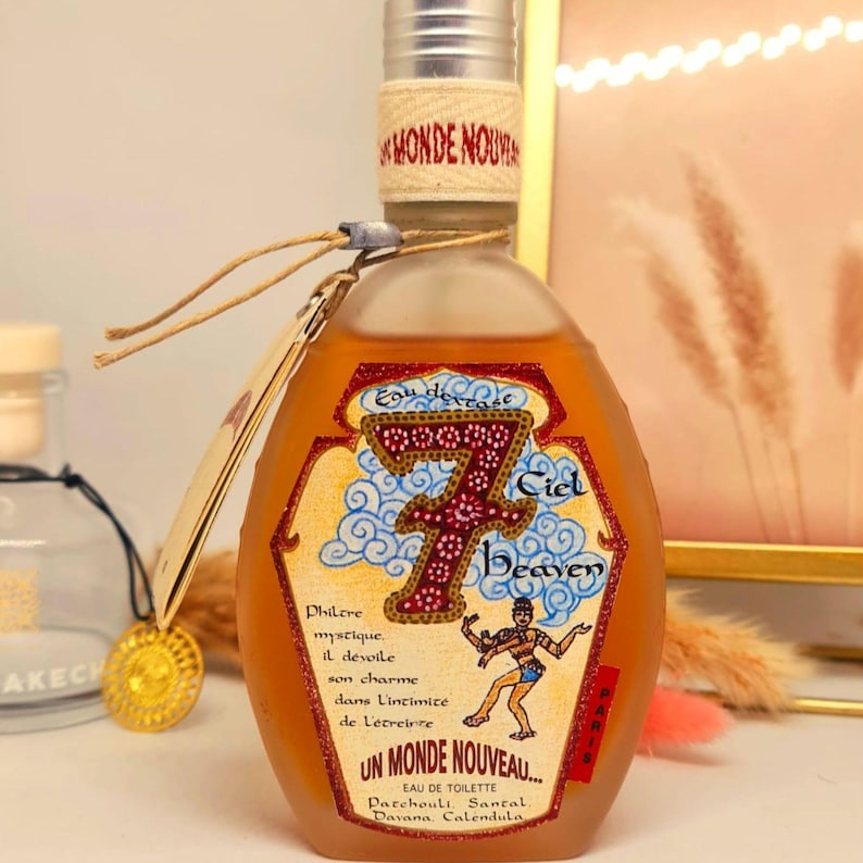 Pu&ograve; includere: Un flacone di profumo con tappo argentato ed etichetta con testo "7 Ciel Heaven" e "Un Monde Nouveau". Il flacone contiene un liquido marrone chiaro e un'etichetta beige con design decorativo.