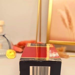 Może przedstawiać: Prostokątne pudełko perfum w kolorze czerwonym i złotym. Na pudełku widnieje napis &bdquo;VAPORISATEUR&rdquo; oraz &bdquo;50 ml e 1.7 FL.OZ&rdquo;. Na spodzie pudełka widoczny jest kod kreskowy. Tło obejmuje złotą ramę i elementy dekoracyjne.