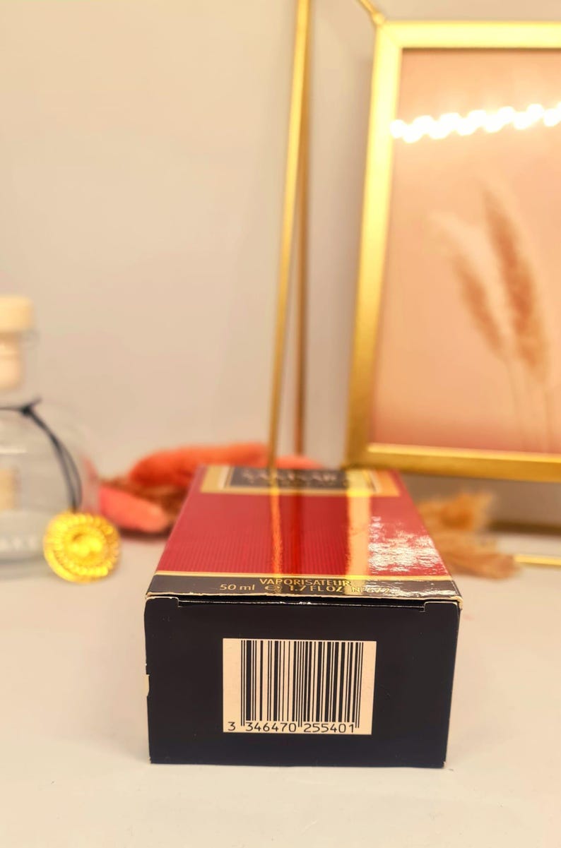 Może przedstawiać: Prostokątne pudełko perfum w kolorze czerwonym i złotym. Na pudełku widnieje napis &bdquo;VAPORISATEUR&rdquo; oraz &bdquo;50 ml e 1.7 FL.OZ&rdquo;. Na spodzie pudełka widoczny jest kod kreskowy. Tło obejmuje złotą ramę i elementy dekoracyjne.