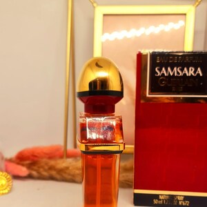 Może przedstawiać: Butelka perfum Samsara Guerlain, ze złotą nakrętką i czerwono-złotym pudełkiem. Butelka perfum jest wypełniona pomarańczowym płynem. Na pudełku widnieje napis "Samsara Guerlain".