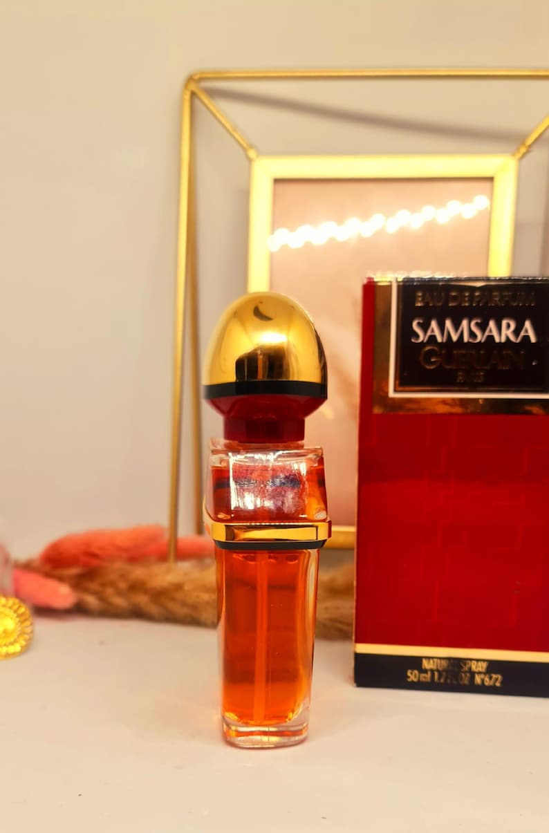 Może przedstawiać: Butelka perfum Samsara Guerlain, ze złotą nakrętką i czerwono-złotym pudełkiem. Butelka perfum jest wypełniona pomarańczowym płynem. Na pudełku widnieje napis "Samsara Guerlain".