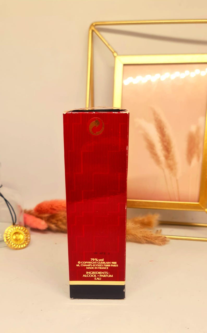 Może przedstawiać: Czerwone, prostokątne pudełko po perfumach ze złotymi akcentami. Na pudełku znajduje się tekst, w tym "79% vol" i "INGREDIENTS: ALCOOL PARFUM EAU". Pudełko znajduje się przed złotą ramką.