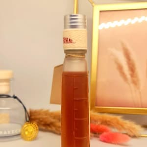 Pu&ograve; includere: Una bottiglia rettangolare trasparente riempita con un liquido marrone, con un tappo argentato e un'etichetta che recita "NUVEAU...". La bottiglia ha segni di misurazione sul lato e un piccolo cartellino. Lo sfondo include una cornice dorata ed elementi decorativi.